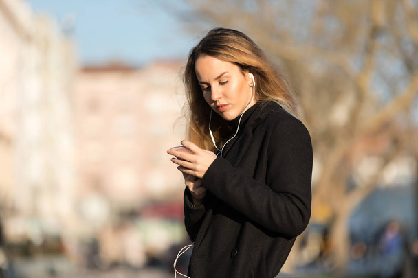 Mooi meisje luisteren naar de muziek met oordopjes van een slimme telefoon met een stedelijke ongerichte achtergrond - Foto, afbeelding