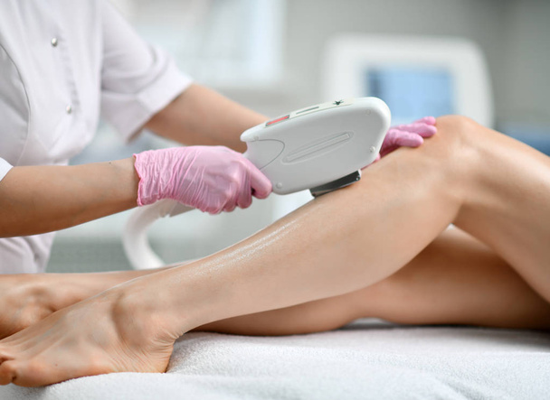 Closeup. Doctor-cosmetician in gloves performes a procedure on patient leg with medical equipment laser in equiped beauty shop - Fotoğraf, Görsel