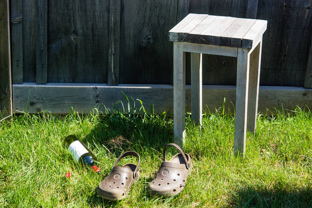 Two old shoes, a stool and an empty bottle of wine on the grass near the fence - Фото, зображення