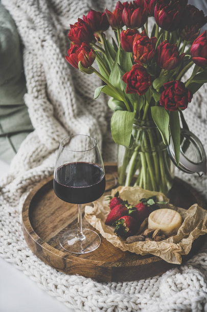Wine snack set with flowers. Glass of red wine, cheese, roasted almonds, strawberries and bouquet of Spring red tulips on wooden tray over white knitted blanket. Romantic mood concept - Foto, Imagen