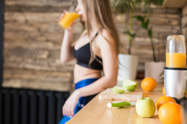 Attractive girl in sportswear prepares a healthy breakfast before the workout in the kitchen, cuts fruit and drinks fresh juice made in a juicer. The benefits of healthy eating, fitness and beauty - 写真・画像