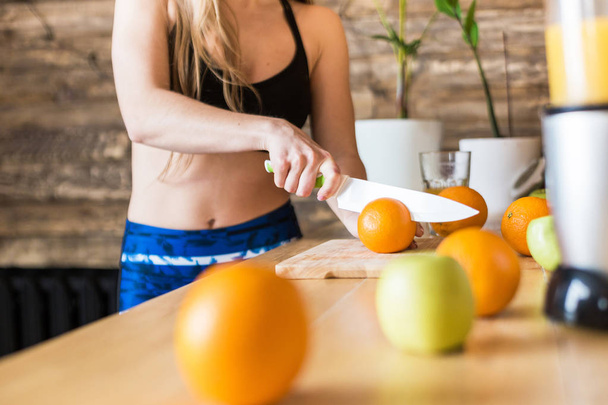 Attractive girl in sportswear preparing a healthy breakfast in the kitchen before training, cutting fruit and making fresh juice. Healthy lifestyle, fitness and beaut - 写真・画像