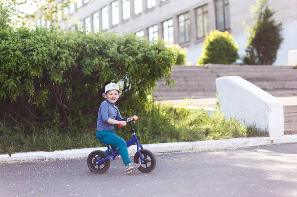 a four-year-old cheerful boy in a hat and sneakers rides a running bike and pushes his feet off the asphalt. - Photo, Image