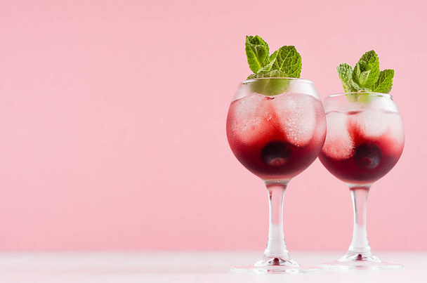 Modern refreshing alcohol red drinks in wineglasses with ice cubes, blueberry, green mint on white wood board and pastel pink background. - Photo, image