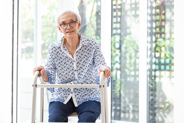 Asiática mujer mayor sentada con andador durante la rehabilitación, anciana mujer usar gafas, sonriendo y mirando a la cámara en casa
 - Foto, Imagen