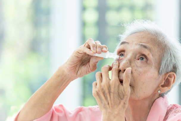 Mulher sênior asiática colocando colírio, visão de close-up da pessoa idosa usando garrafa de colírio nos olhos, mulher idosa doente que sofre de olho irritado, sintomas ópticos, conceito de saúde
 - Foto, Imagem
