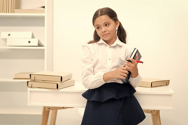 Ready for lesson. Girl child holds book while stand near table white background. Schoolgirl enjoy studying and reading books on lessons. Kid school uniform smiling face likes visiting school lessons - Фото, изображение