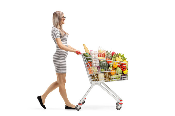 Full length profile shot of a young blond woman pushing a shopping cart with food products and walking isolated on white background - Foto, immagini