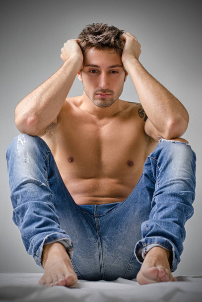 Depressed or sad handsome young man shirtless in jeans, sitting on floor with head in his hands - Fotoğraf, Görsel