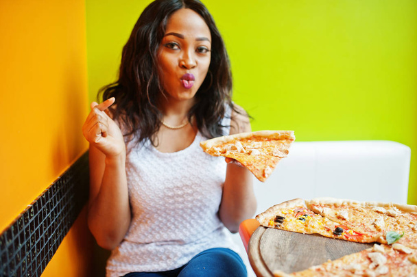 明るい色のレストランに座ってピザを持つアフリカの女性. - 写真・画像