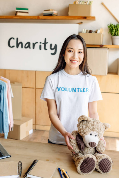 симпатичная азиатская девушка-волонтер держит плюшевого мишку, улыбается и смотрит в камеру
 - Фото, изображение