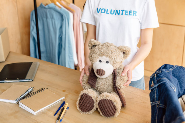 cropped view of volunteer girl holding teddy bear while standing near wooden table with notebooks  - Фото, изображение