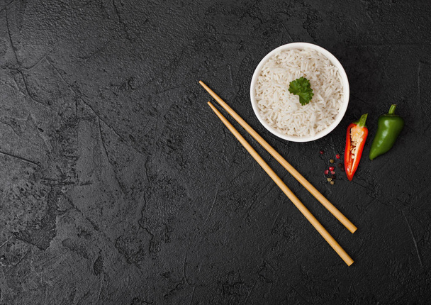 White bowl with boiled organic basmati jasmine rice with black chopsticks on black stone background with green and red pepper. Space for text - Foto, Bild