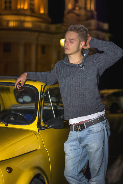 Night shot of handsome blond young man standing next to small yellow car - Foto, afbeelding