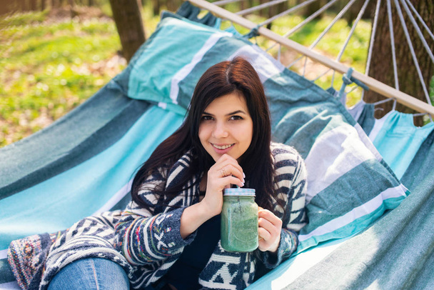 Feliz chica comiendo sano bebiendo licuado verde desintoxicación al aire libre en hamaca. Mujer en dieta de pérdida de peso nutrición vegana limpiar
. - Foto, Imagen