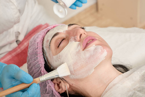 Young woman in beauty salon does make application of moisturizing, softening, regenerating mask. - Photo, Image