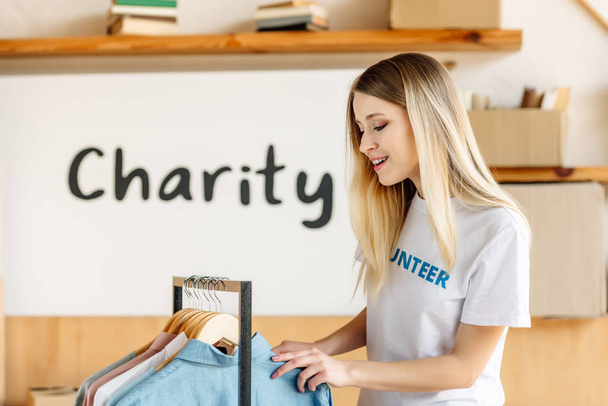 pretty blonde volunteer standing near rack with different shirts - Foto, Bild