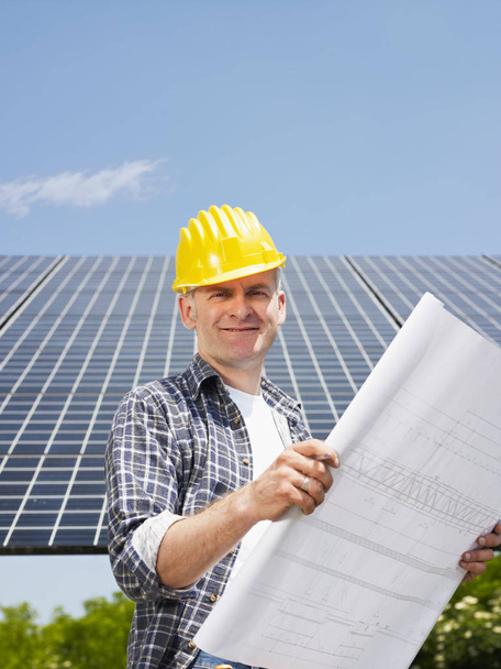Portrait of mid adult italian male electrician reading blueprints in solar power station and smiling. Vertical shape, side view. Copy space - Foto, imagen
