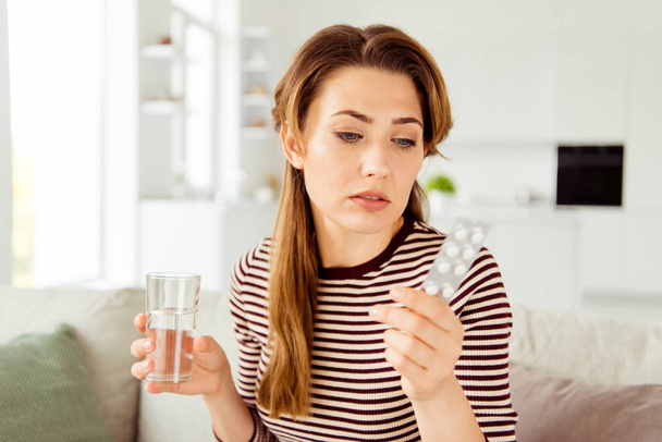 Close up photo of serious lady contemplate discomfort hurt injury hold medicine pills cure virus allergy inflammation have striped shirt long curly wavy hair wear sit divan in light apartment - Foto, Bild