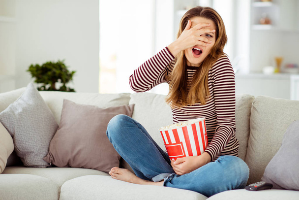 Close up photo beautiful she her model lady hand arm hide face awful ugh scene scary movie big container corns wear jeans denim striped pullover clothes sit comfort divan house living room indoors - 写真・画像