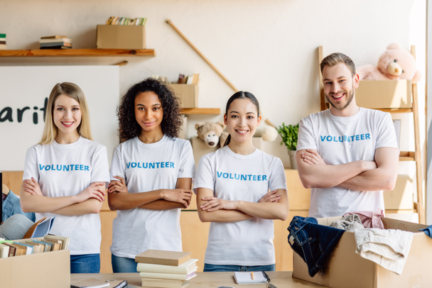 четыре улыбающихся молодых волонтера в белых футболках с волонтерскими надписями, улыбающимися и смотрящими в камеру
 - Фото, изображение