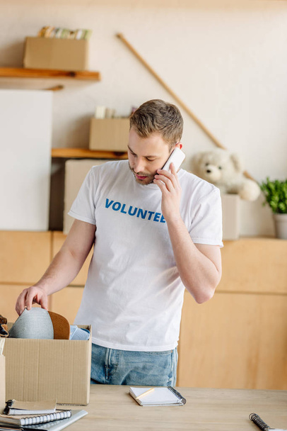 handsome volunteer talking on smartphone while standing near carton box with clothes - Foto, Bild