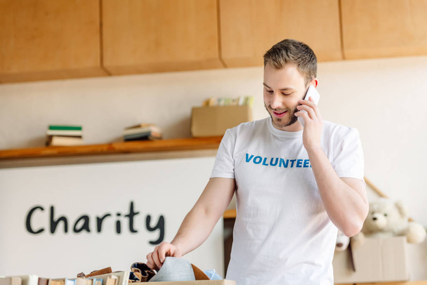 handsome young volunteer talking on smartphone while standing near cardboard box with clothes - Photo, Image