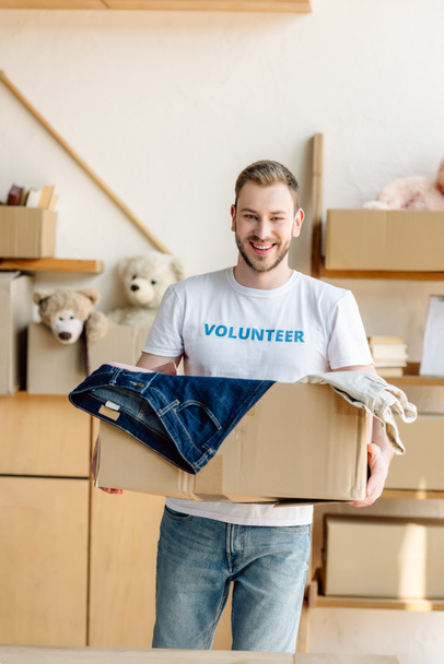 handsome, cheerful volunteer holding cardboard box with clothes - Zdjęcie, obraz