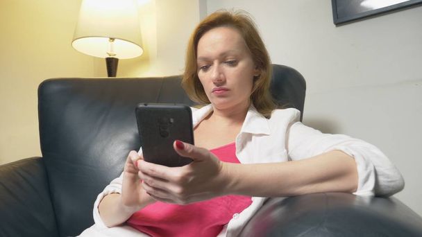A woman is using a smartphone at home, sitting in a large leather chair in the evening, in the background a floor lamp - Foto, Imagem