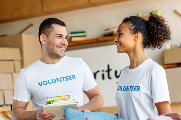 two cheerful multicultural volunteers talking while holding books and clothes - Foto, afbeelding