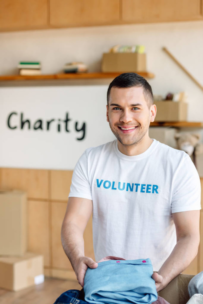 guapo, voluntario sonriente sosteniendo la ropa y mirando a la cámara en el centro de caridad
 - Foto, imagen