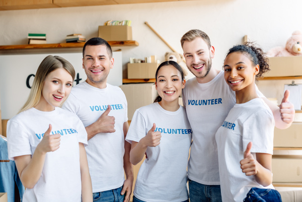 cinco jóvenes voluntarios multiculturales mostrando los pulgares hacia arriba, sonriendo y mirando a la cámara
 - Foto, Imagen