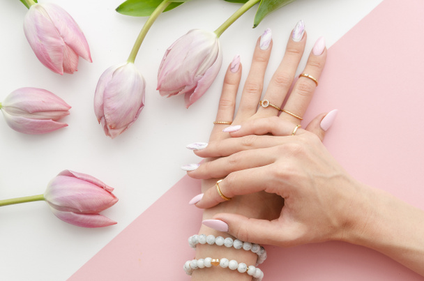 Top θέα γυναικεία χέρια με ένα μανικιούρ και το σχέδιο πάνω στα νύχια. Ρύθμιση με ροζ τουλίπα λουλούδια και πέρλες σε λευκό και ροζ φόντο για Φέιγ βολάν σαλονιού ομορφιάς - Φωτογραφία, εικόνα