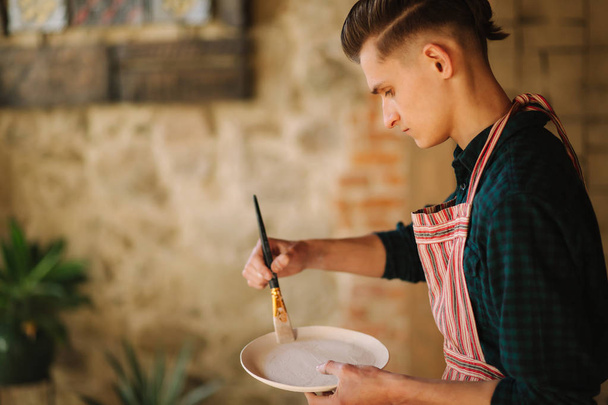 Fiatal művész díszt készít kerámia tányéron. Jóképű fiatalember, a műhelyében dolgozik. - Fotó, kép