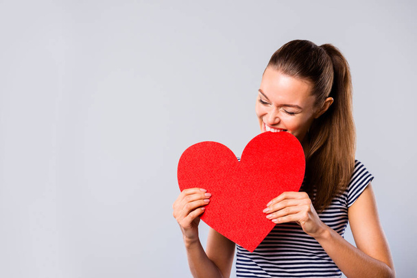 Close up photo beautiful amazing she her lady hold hands arms big large red paper heart shape figure postcard bite it laughter game play wear blue white striped t-shirt isolated grey background - Foto, imagen