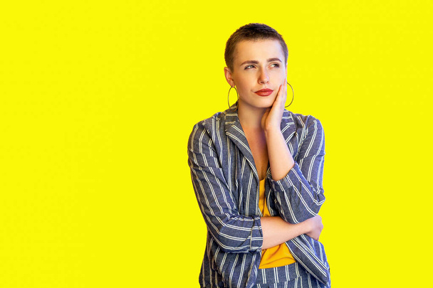 thoughtful young short hair woman in yellow shirt and striped suit touching her face and looking aside while posing on yellow background - Photo, image