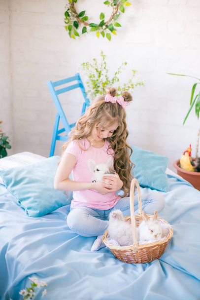 Mała śliczna dziewczyna z długimi kręconymi włosami na łóżku z małymi króliczkami i wielkanocnym wystrojem w domu. - Zdjęcie, obraz