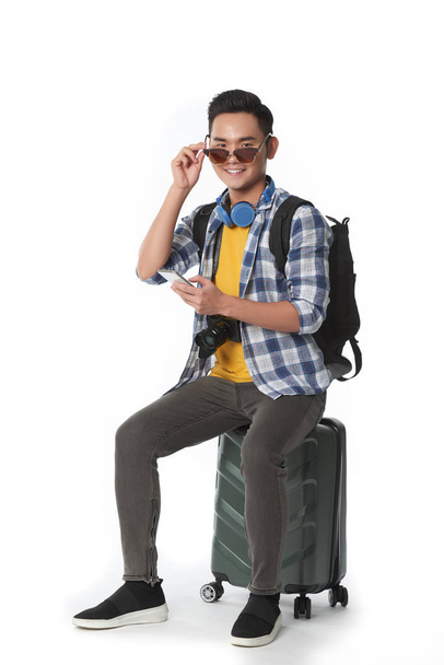 Smiling happy traveler sitting on suitcase and putting on sunglasses - Foto, Imagem