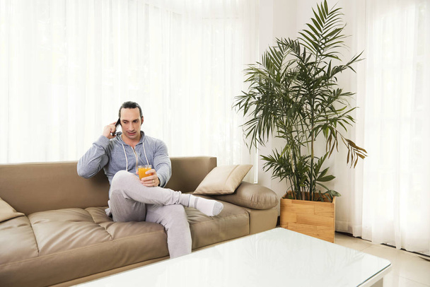 Serious man in comfortable clothes drinking fresh juice and calling on phone - Foto, Imagen