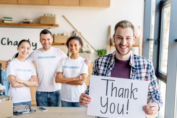 selective focus of handsome man holding placard with thank you text while standing near multicultural volunteers in charity center - Photo, Image