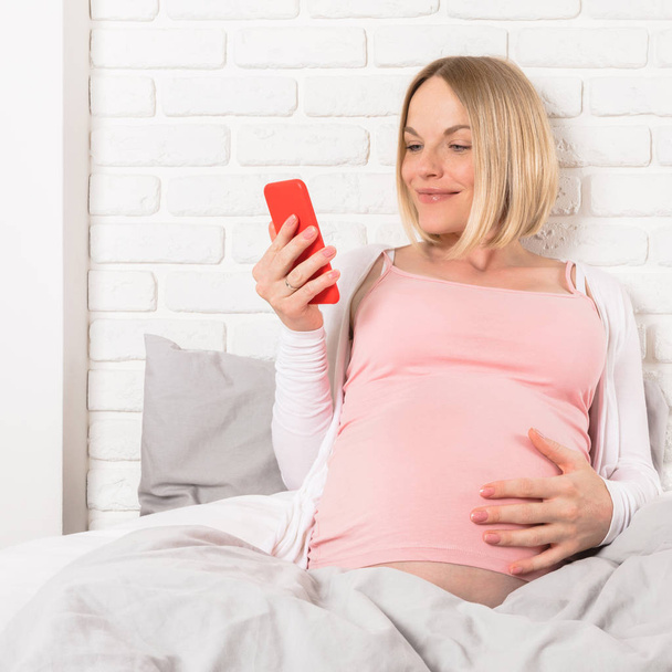 Szczęśliwy uśmiechający się kobieta w ciąży leży w łóżku i patrzy na telefon komórkowy - Zdjęcie, obraz
