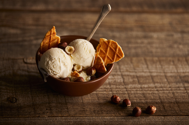 delicious ice cream with pieces of waffle, spoon and hazelnuts in bowl on wooden surface - Photo, image