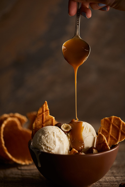 Cropped view of woman pouring caramel on ice cream with pieces of waffle and hazelnuts - Foto, afbeelding