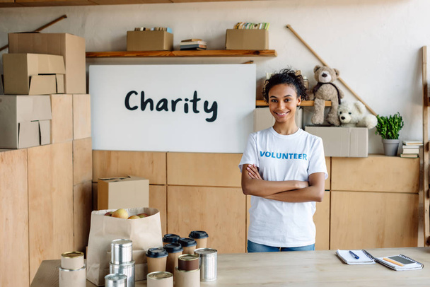 selektivní zaměření usmívající se africké americké dobrovolnice stojící poblíž stolu s konzervovanými potravinami a papírovými šálky - Fotografie, Obrázek
