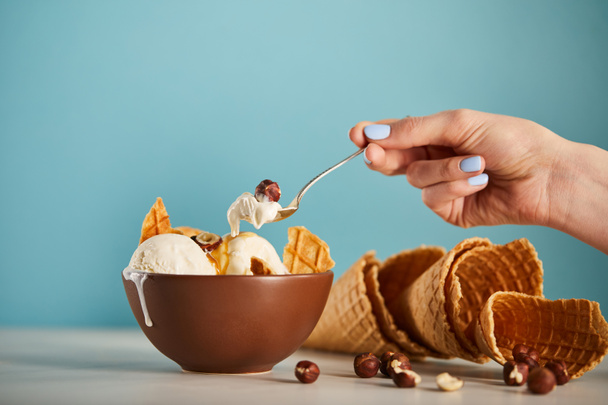cropped view of woman with spoon, bowl of ice cream, hazelnuts and waffle cones on blue - Foto, imagen