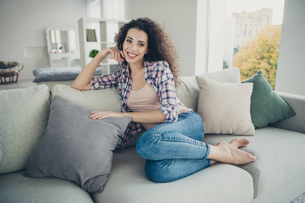 Portrait of attractive pretty teen teenager sit divan student glad content have vacation touch pillow wear modern spring trendy checked denim outfit bare foot in living room - Foto, Imagem