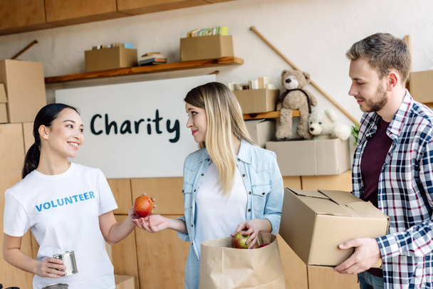 lachende Aziatische vrijwilliger geeft ingeblikt voedsel en appels aan man en vrouw in liefdadigheids centrum - Foto, afbeelding