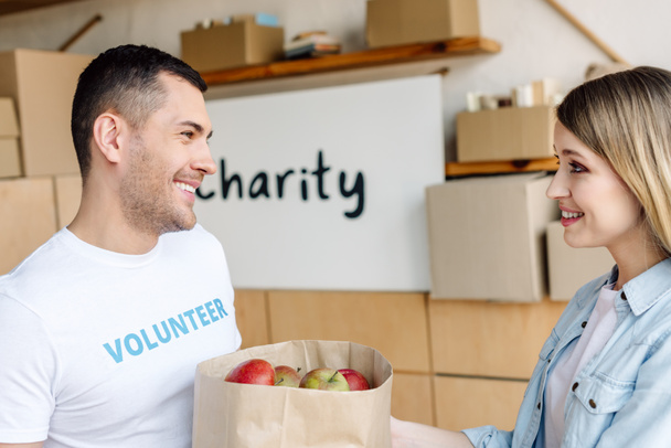knappe, lachende vrijwilliger geven papieren zak met appels aan aantrekkelijke vrouw - Foto, afbeelding