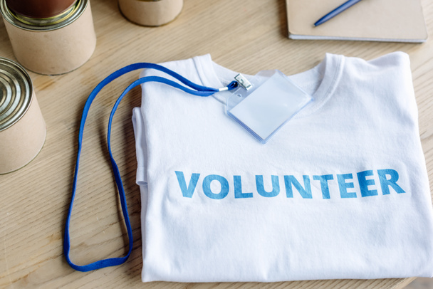 fehér póló kék önkéntes felirattal, jelvénnyel és konzervdobozzal a fa asztalon - Fotó, kép