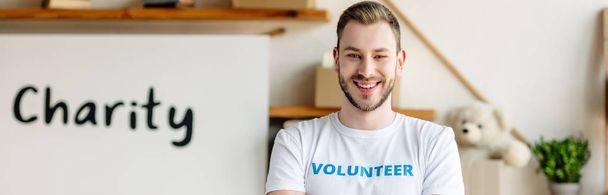 Panoramaaufnahme eines gut aussehenden, lächelnden Freiwilligen, der neben einem Plakat mit Charity-Aufschrift steht - Foto, Bild